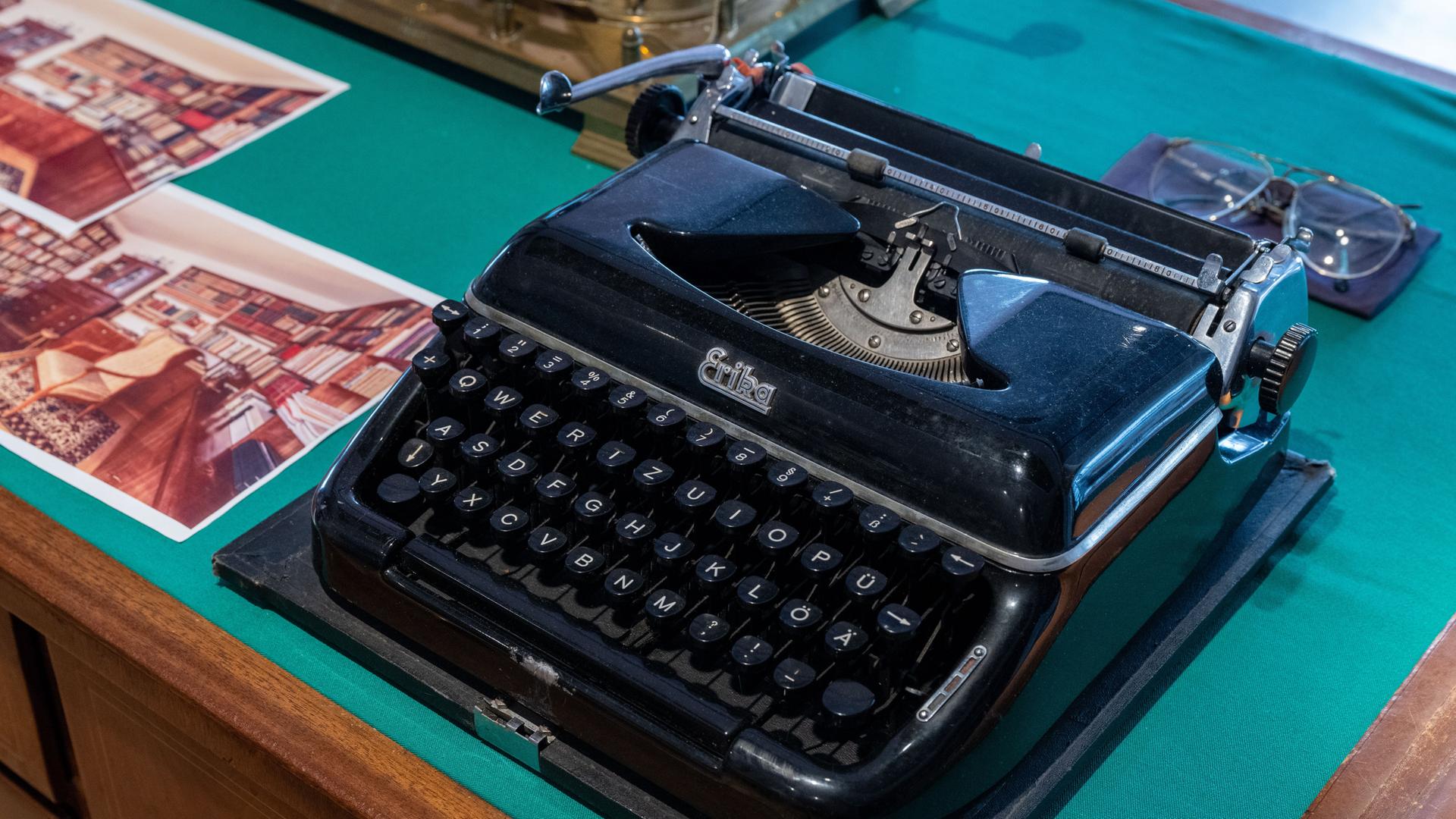 Eine schwarze Schreibmaschine auf einem Tisch neben einem Foto, welches ein Zimmer mit vielen Büchern zeigt. Sie gehörte dem Schriftsteller Stefan Heym und ist in der Arbeitsbibliothek Heyms im Kulturzentrum in Chemnitz ausgestellt. 