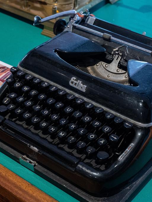 Eine schwarze Schreibmaschine auf einem Tisch neben einem Foto, welches ein Zimmer mit vielen Büchern zeigt. Sie gehörte dem Schriftsteller Stefan Heym und ist in der Arbeitsbibliothek Heyms im Kulturzentrum in Chemnitz ausgestellt. 