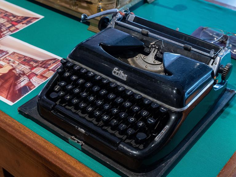 Eine schwarze Schreibmaschine auf einem Tisch neben einem Foto, welches ein Zimmer mit vielen Büchern zeigt. Sie gehörte dem Schriftsteller Stefan Heym und ist in der Arbeitsbibliothek Heyms im Kulturzentrum in Chemnitz ausgestellt. 