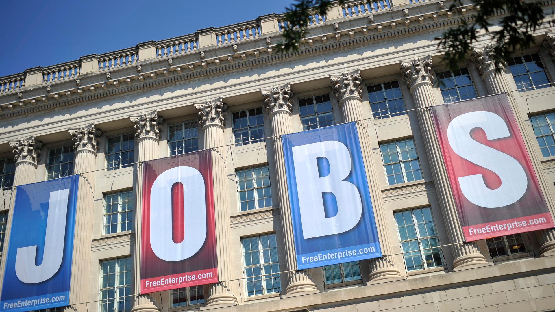 Blick auf die Vorderfront des klassizistischen Gebäudes, die man nicht vollständig sieht. Daran hängen vier rote beziehungsweise blaue Transparente mit jeweils einem Buchstaben, die zusammen das Wort "Jobs" ergeben. 