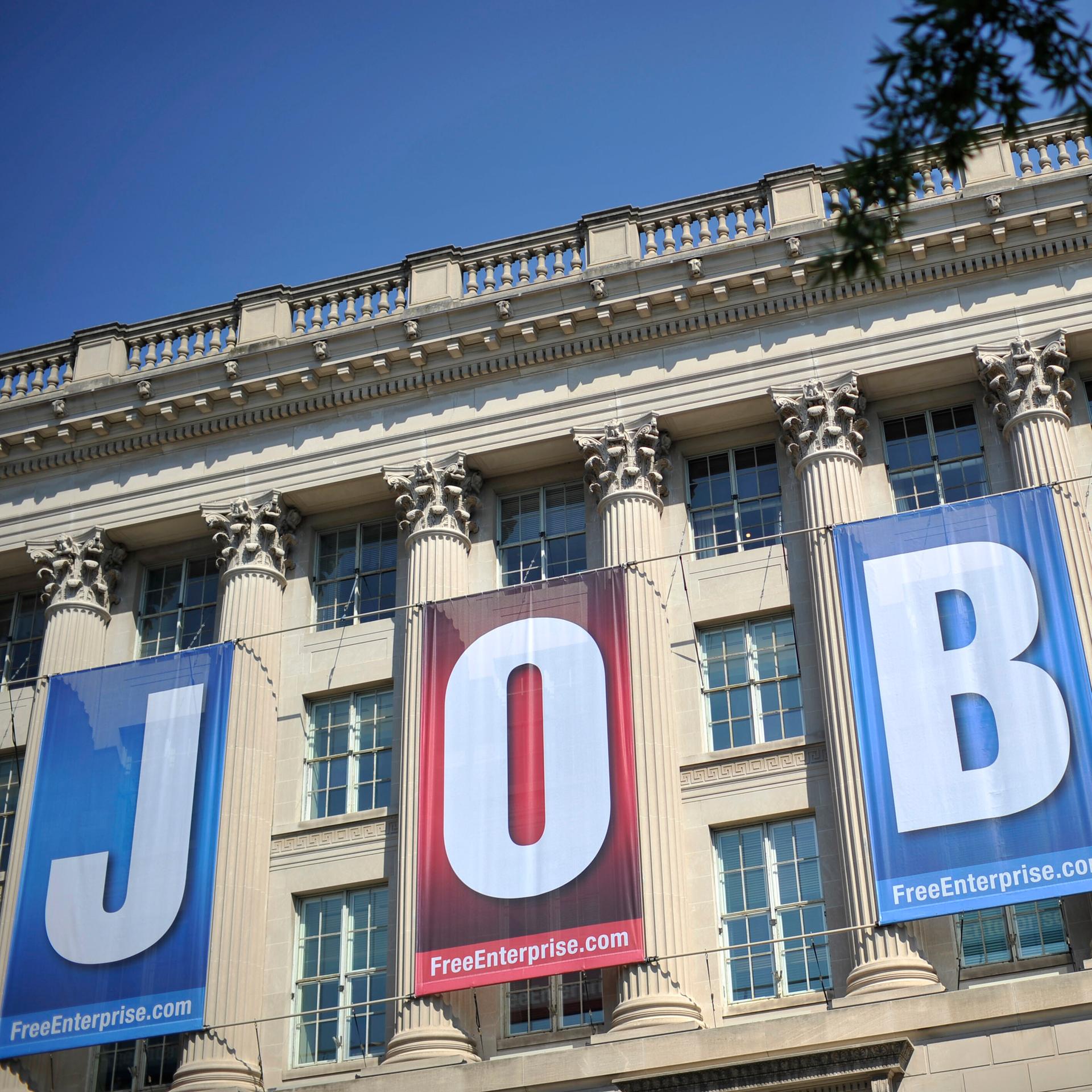 Blick auf die Vorderfront des klassizistischen Gebäudes, die man nicht vollständig sieht. Daran hängen vier rote beziehungsweise blaue Transparente mit jeweils einem Buchstaben, die zusammen das Wort "Jobs" ergeben. 