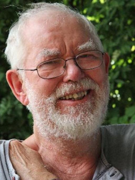 Der Komponist in einer grünen Umgebung mit weißem Haar und weißem Bart lacht in die Kamera.