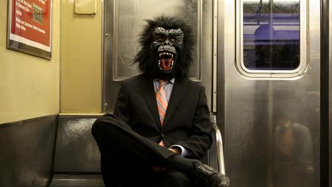 Ein Mann im schwarzen Anzug und mit Gorillamaske sitzt in einer U-Bahn.