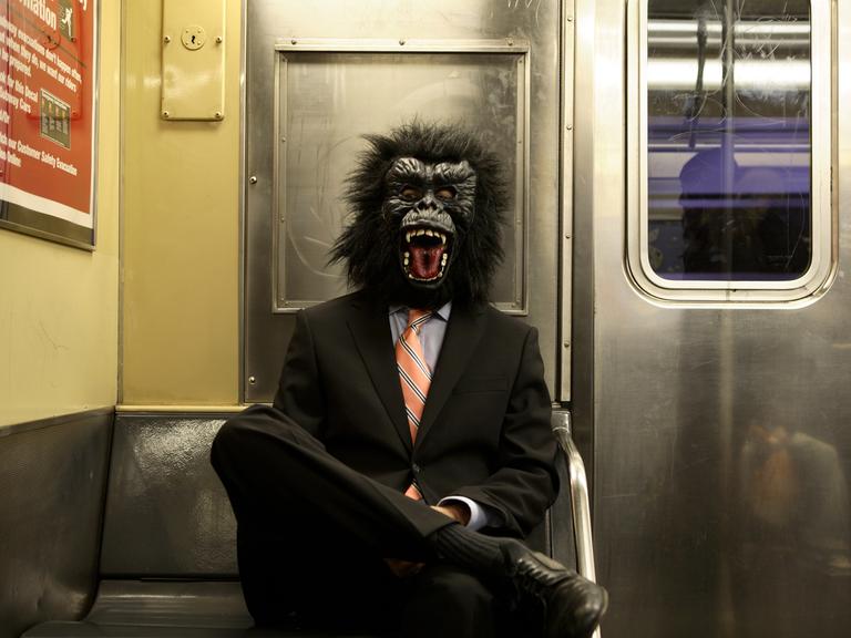 Ein Mann im schwarzen Anzug und mit Gorillamaske sitzt in einer U-Bahn.