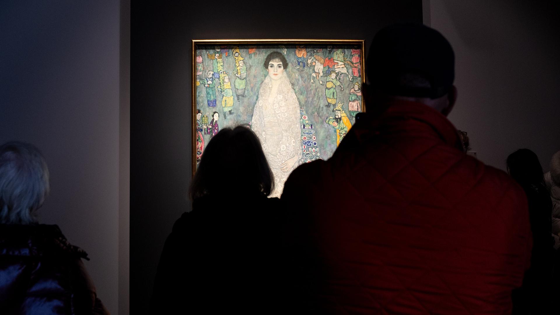 Gäste im Auktionshaus Sotheby's betrachten das Gemälde von Gustav Klimt, das in einem abgedunkelten Raum beleuchtet vor ihnen hängt.