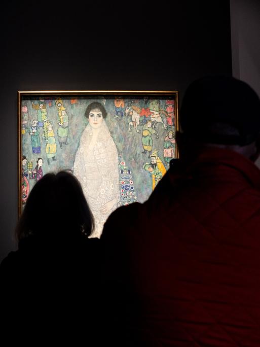 Gäste im Auktionshaus Sotheby's betrachten das Gemälde von Gustav Klimt, das in einem abgedunkelten Raum beleuchtet vor ihnen hängt.