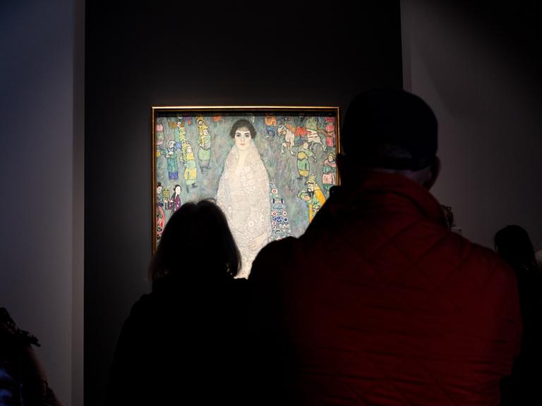Gäste im Auktionshaus Sotheby's betrachten das Gemälde von Gustav Klimt, das in einem abgedunkelten Raum beleuchtet vor ihnen hängt.