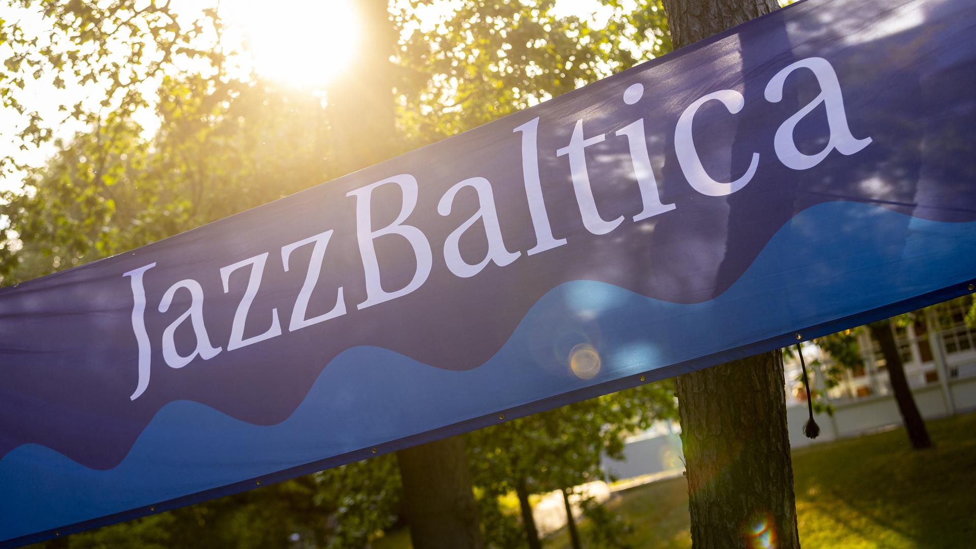 Ein Banner mit dem Schriftzug JazBaltica und einem Wellenmotiv ist zwischen Bäumen gespannt, durch die die Sonne hindurchlugt.
