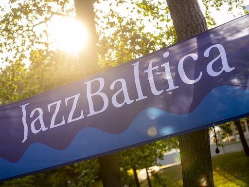 Ein Banner mit dem Schriftzug JazBaltica und einem Wellenmotiv ist zwischen Bäumen gespannt, durch die die Sonne hindurchlugt.