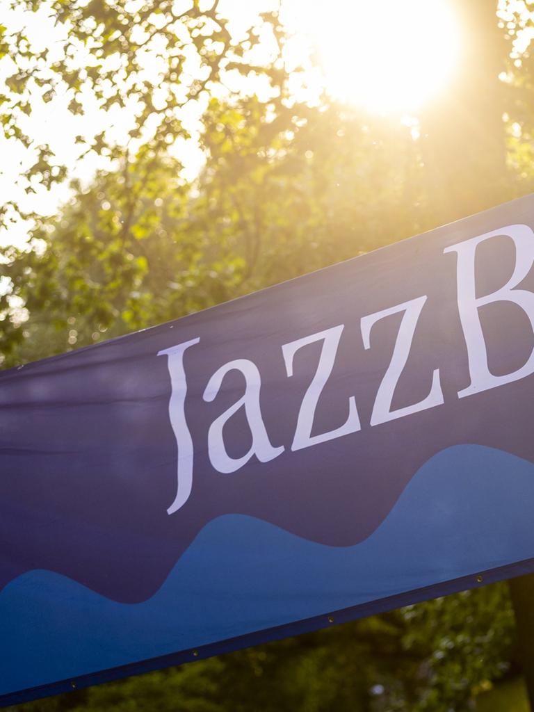 Ein Banner mit dem Schriftzug JazBaltica und einem Wellenmotiv ist zwischen Bäumen gespannt, durch die die Sonne hindurchlugt.