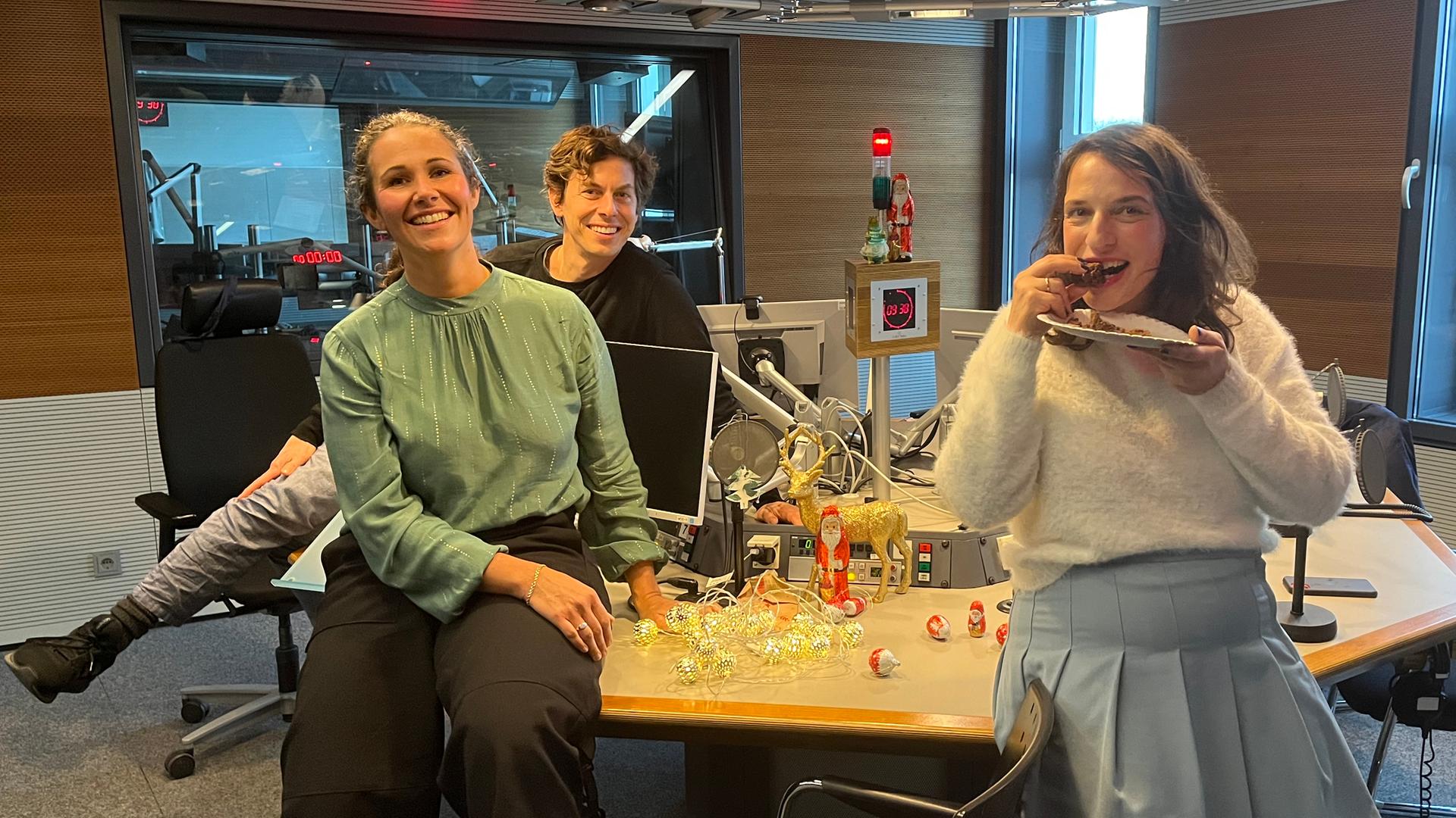 Sonja Koppitz, Utz Dräger und Caro Korneli sitzen auf dem Studiotisch. In der Mitte stehen Bildschirme und Mikrofone. Caro Korneli beißt in ein Stück Kuchen.