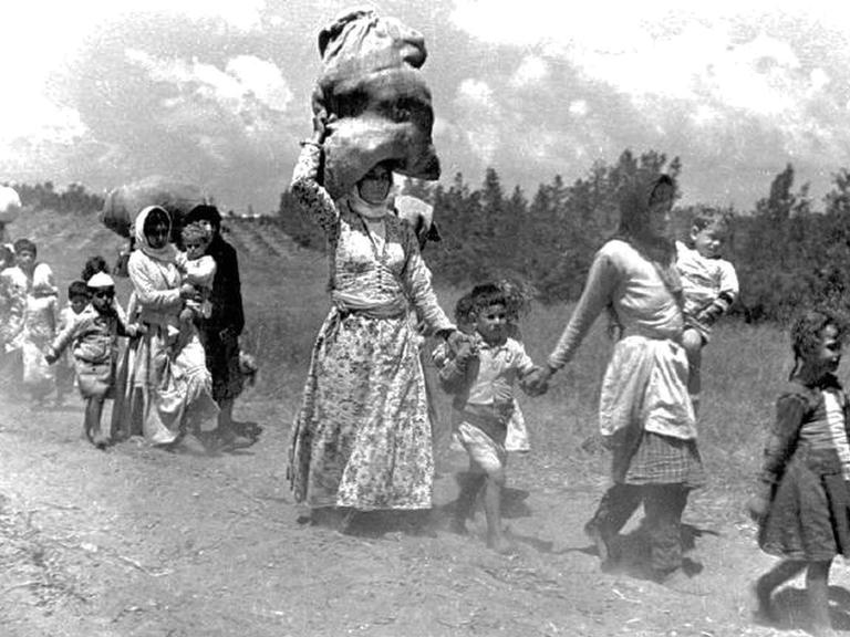 1948: Palästinensische Frauen und Kinder auf der Flucht. Eine Fraut trägt einen großen Sack auf ihrem Kopf.