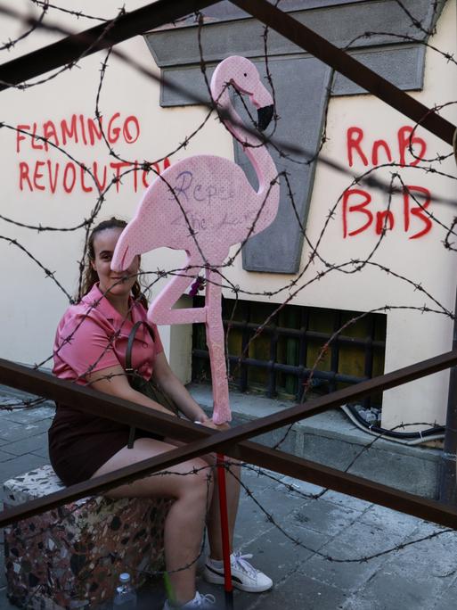 Ein Mitglied der sogenannten „Flamingo-Revolution“ sitzt mit einer symbolischen Flamingo-Skulptur hinter einer Stacheldrahtinstallation.