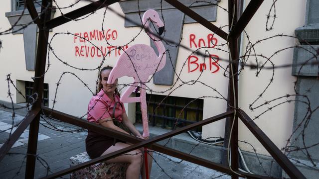 Ein Mitglied der sogenannten „Flamingo-Revolution“ sitzt mit einer symbolischen Flamingo-Skulptur hinter einer Stacheldrahtinstallation.