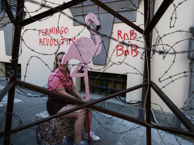 Ein Mitglied der sogenannten „Flamingo-Revolution“ sitzt mit einer symbolischen Flamingo-Skulptur hinter einer Stacheldrahtinstallation.