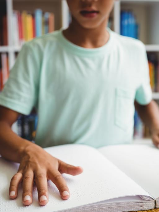 Ein Junge liest ein Buch in Braille-Schrift vor eine Bücherwand. 