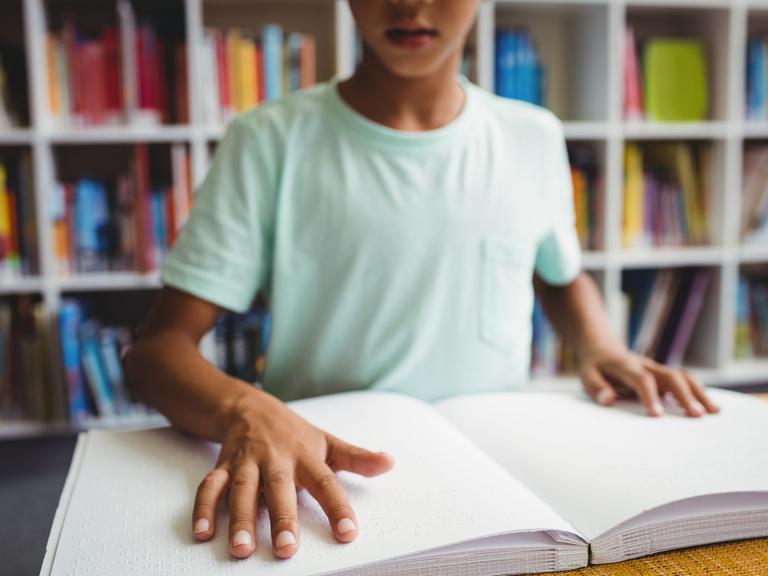 Ein Junge liest ein Buch in Braille-Schrift vor eine Bücherwand. 
