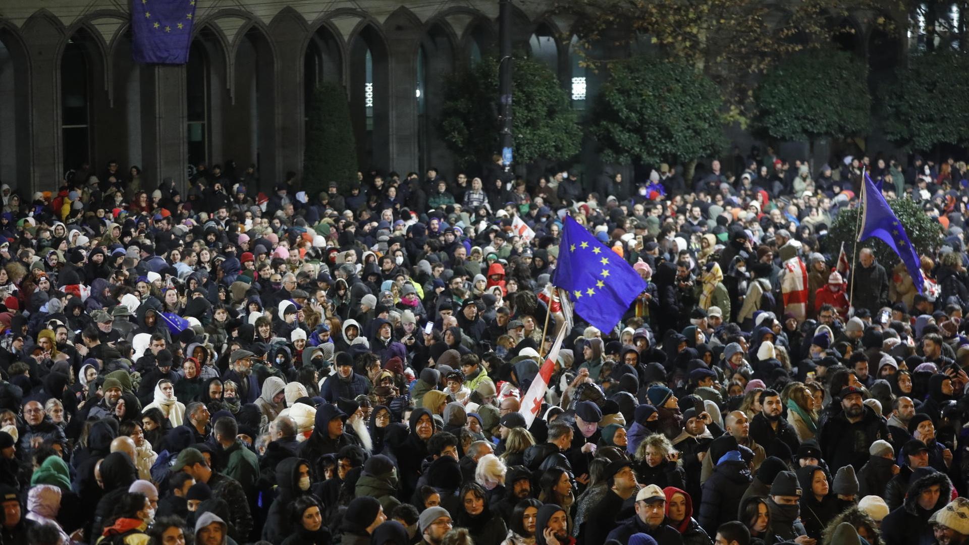 Vor dem georgischen Parlament steht bei Nacht eine Menschenmenge. Einige tragen EU-Flaggen oder georgische Flaggen bei sich.