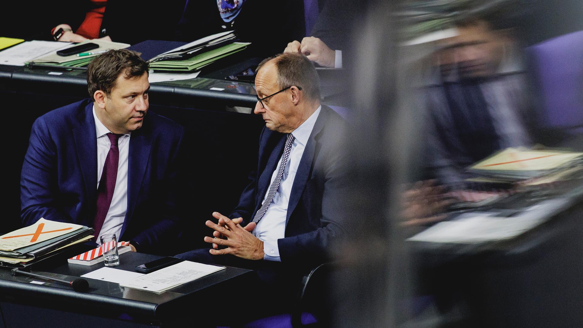 SPD-Chef und Finanzminister Lars Klingbeil (SPD) und Kanzler Friedrich Merz (CDU) sitzen im Bundestag nebeneinander und führen ein ernstes Gespräch. Der Kanzler verzieht dabei das Gesicht. 
