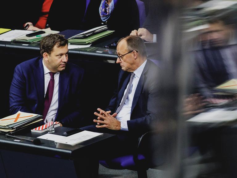 SPD-Chef und Finanzminister Lars Klingbeil (SPD) und Kanzler Friedrich Merz (CDU) sitzen im Bundestag nebeneinander und führen ein ernstes Gespräch. Der Kanzler verzieht dabei das Gesicht. 