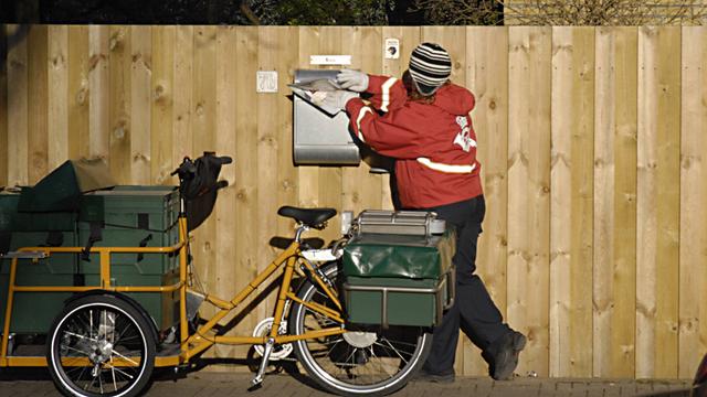 Ein Briefträger wirft Post in einen Briefkasten an einem Haus. Neben ihm steht ein Fahrrad.
