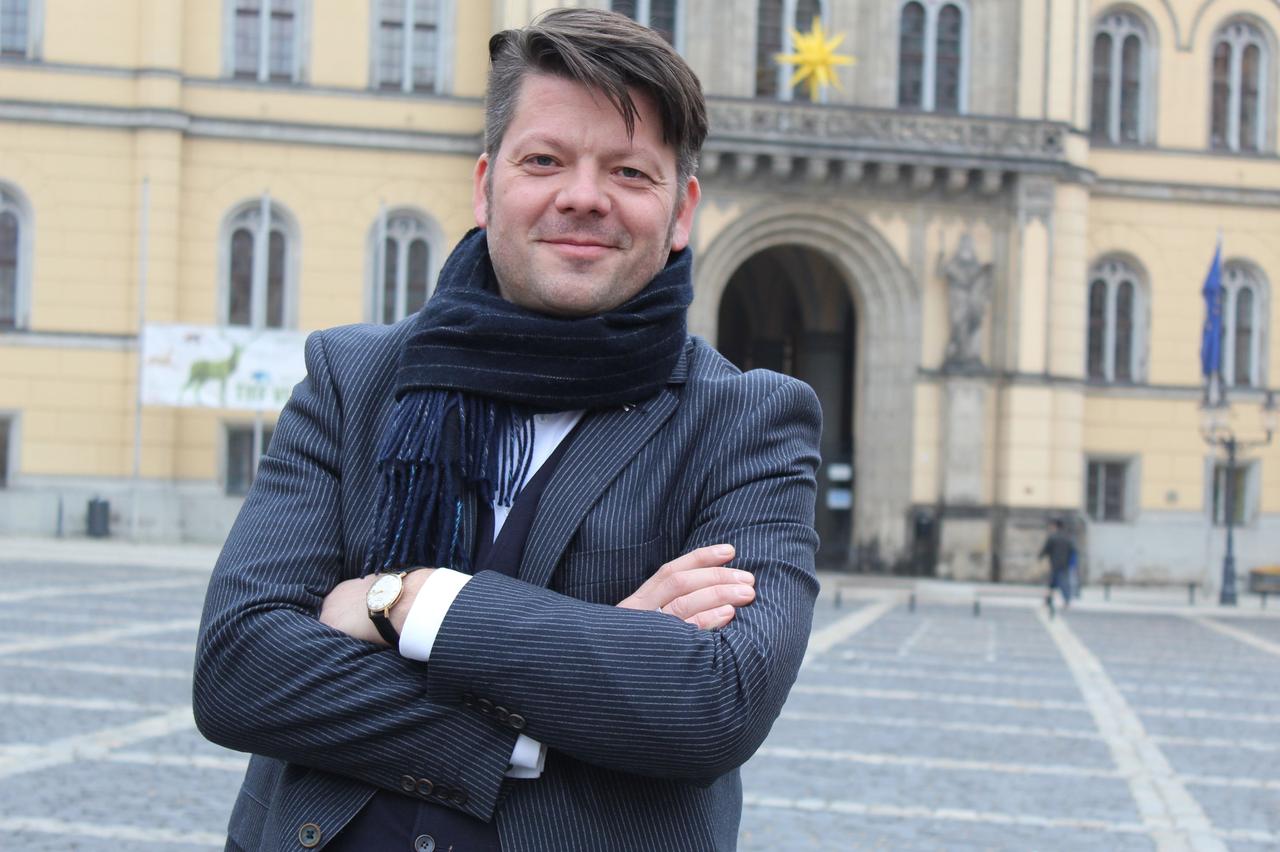 Thomas Zenker, der Oberbürgermeister von Zittau, steht auf einem Platz in Zittau. Er trägt einen dunklen, längsgestreiften Anzug. Der Mittvierziger trägt die dunklen  Haare schwungvoll nach links gekämmt. 