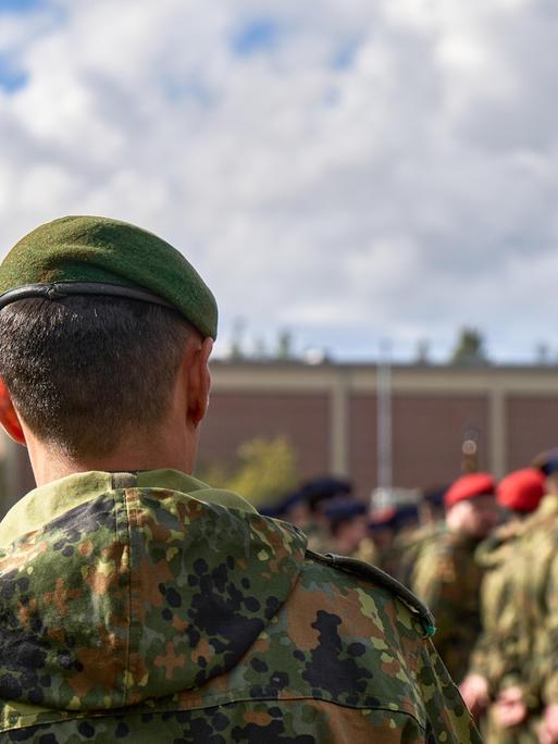 Ein Soldat der Bundeswehr mit grünem Barett steht in Tarnuniform. Weitere Soldaten mit roten Baretts stehen in Reih und Glied verschwommen im Hintergrund