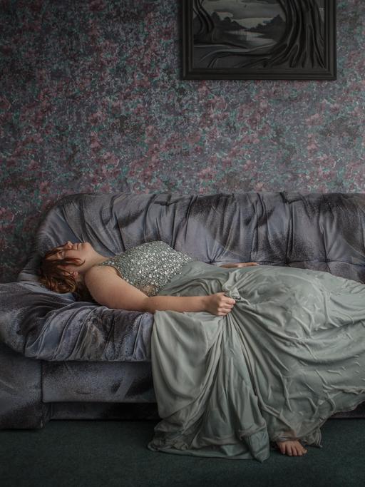 Eine Frau liegt barfuß auf einer Couch und schaut an die Decke. Sie trägt ein hellgrünes Kleid. 