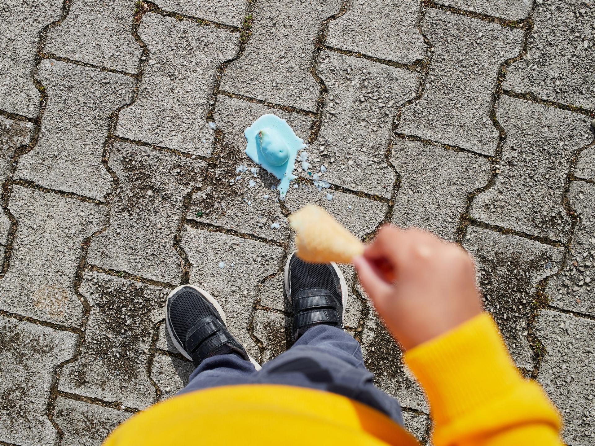 Blick vor die Füße einer Person. Dort liegt eine schmelzende Eiskugel, die von der Waffel gefallen ist.