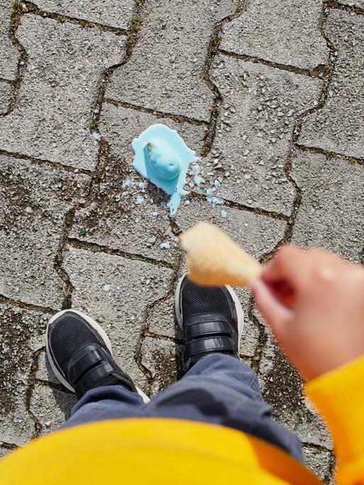 Blick vor die Füße einer Person. Dort liegt eine schmelzende Eiskugel, die von der Waffel gefallen ist.