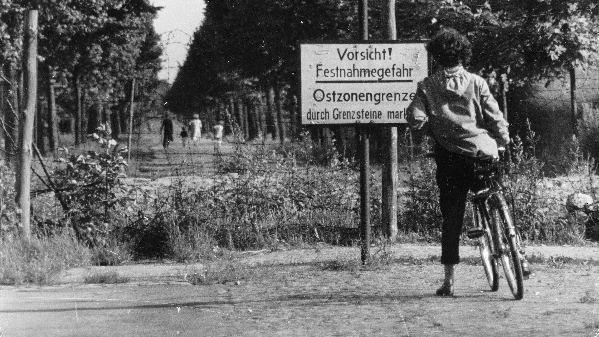 Eine Frau mit Fahrrad nähert sich einem unbefestigten Weg an der Grenze zwischen Westsektor und Ostzone in Berlin. Zwei Schilder warnen: "Achtung! hier endet der Westsektor und Vorsicht!"  Eine Frau mit Fahrrad nähert sich einem unbefestigten Weg an der Grenze zwischen Westsektor und Ostzone in Berlin. Zwei Schilder warnen: "Achtung! hier endet der Westsektor und Vorsicht!"