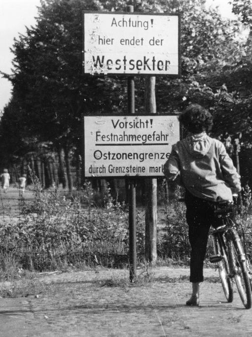 Eine Frau mit Fahrrad nähert sich einem unbefestigten Weg an der Grenze zwischen Westsektor und Ostzone in Berlin. Zwei Schilder warnen: "Achtung! hier endet der Westsektor und Vorsicht!"  Eine Frau mit Fahrrad nähert sich einem unbefestigten Weg an der Grenze zwischen Westsektor und Ostzone in Berlin. Zwei Schilder warnen: "Achtung! hier endet der Westsektor und Vorsicht!"