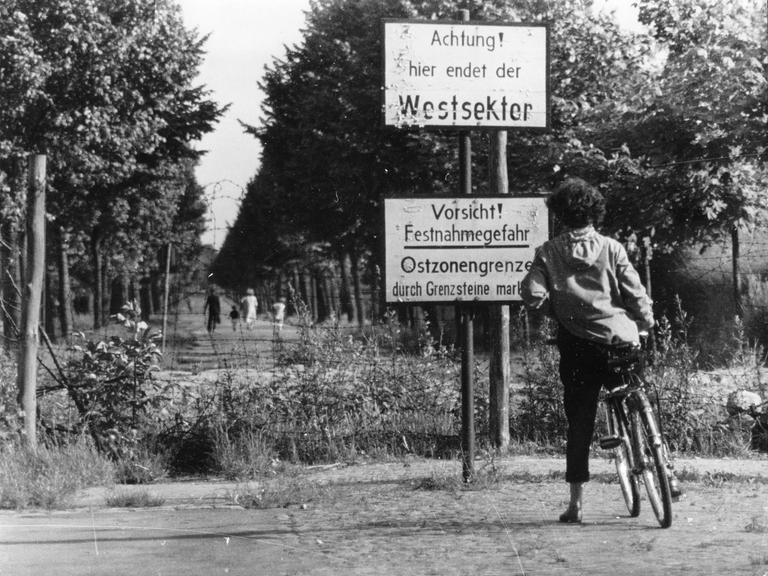 Eine Frau mit Fahrrad nähert sich einem unbefestigten Weg an der Grenze zwischen Westsektor und Ostzone in Berlin. Zwei Schilder warnen: "Achtung! hier endet der Westsektor und Vorsicht!"  Eine Frau mit Fahrrad nähert sich einem unbefestigten Weg an der Grenze zwischen Westsektor und Ostzone in Berlin. Zwei Schilder warnen: "Achtung! hier endet der Westsektor und Vorsicht!"