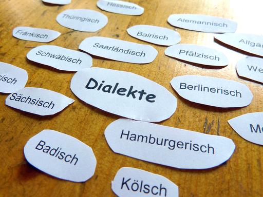 Papierausschnitte mit den Namen mehrerer deutscher Dialekte liegen auf einem Holztisch.
