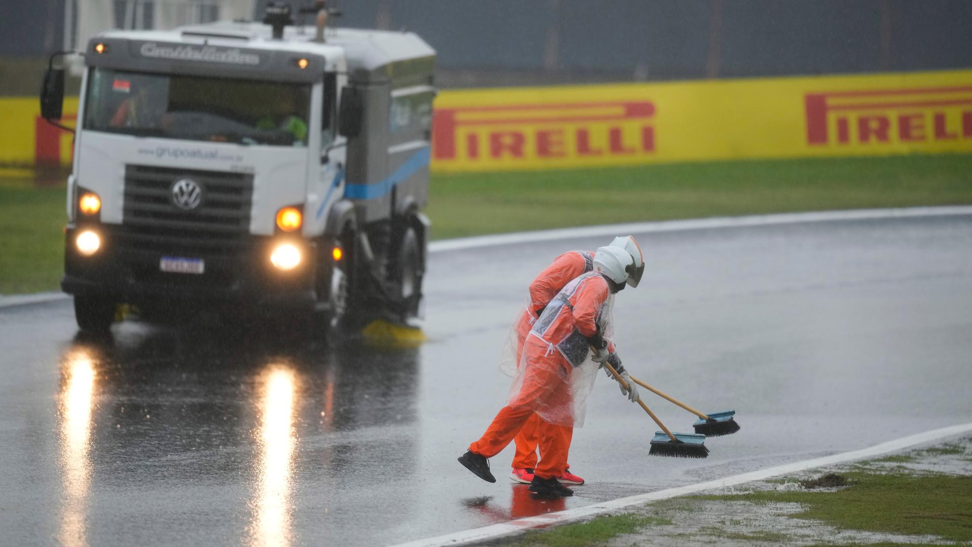 Zwei Männer in orangefarbenen Overalls und mit Helmen versuchen, mit Besen das Wasser von einer Rennstrecke zu entfernen. Sie tragen einen Regenschutz. 