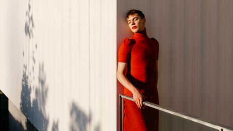 Ein Transgender Fluid Mann im roten Kleid und rotem Lippenstift steht im Sonnenlicht an eine Wand gelehnt.
