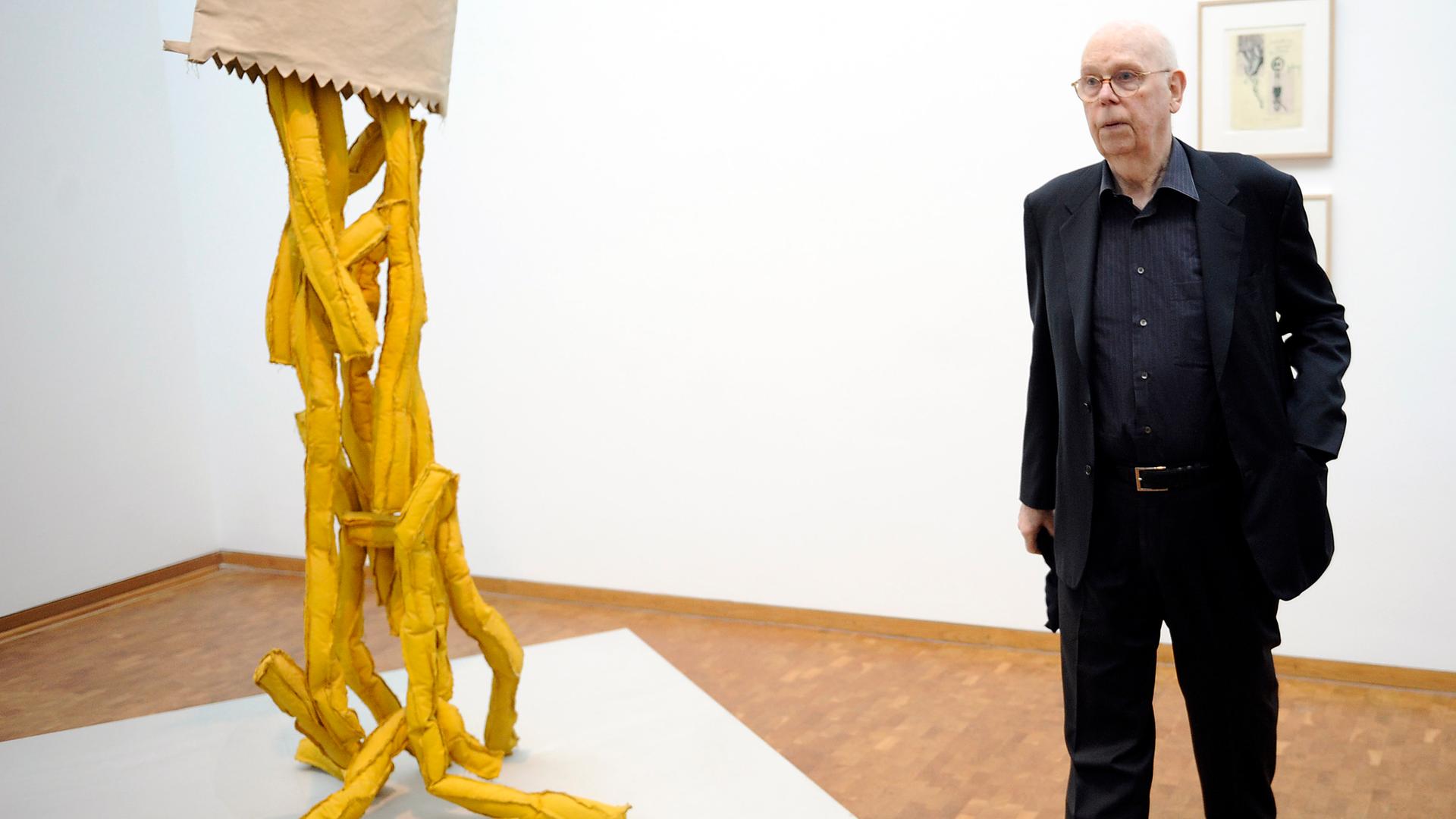Der Künstler Claes Oldenburg steht neben einer überdimensionalen, nach unten geöffneten Tüte Pommes Frites, eine seiner prägnanten Skulpturen bei einer Ausstellung in Köln im Jahr 2012.