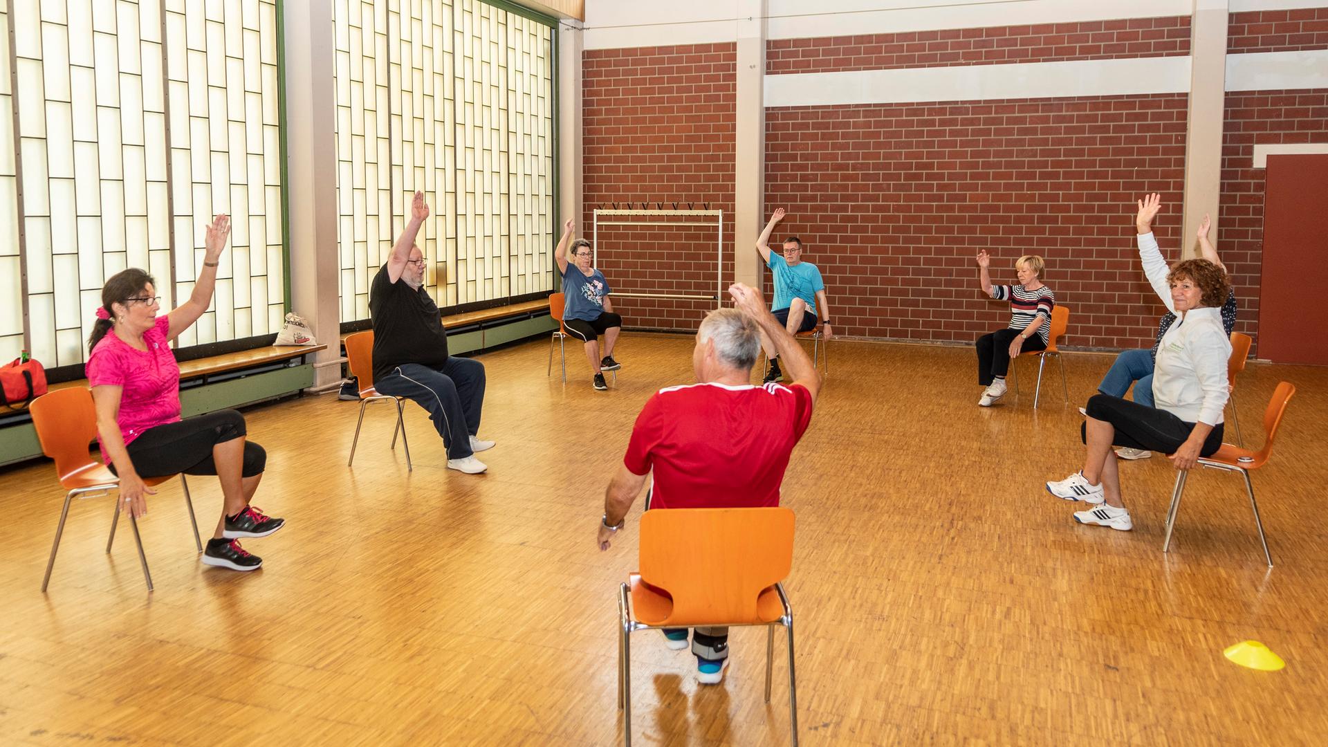 Senioren sitzen  im Rahmen eines Präventionskurses in einer Sporthalle auf Stühlen und strecken einen Arm nach oben. 