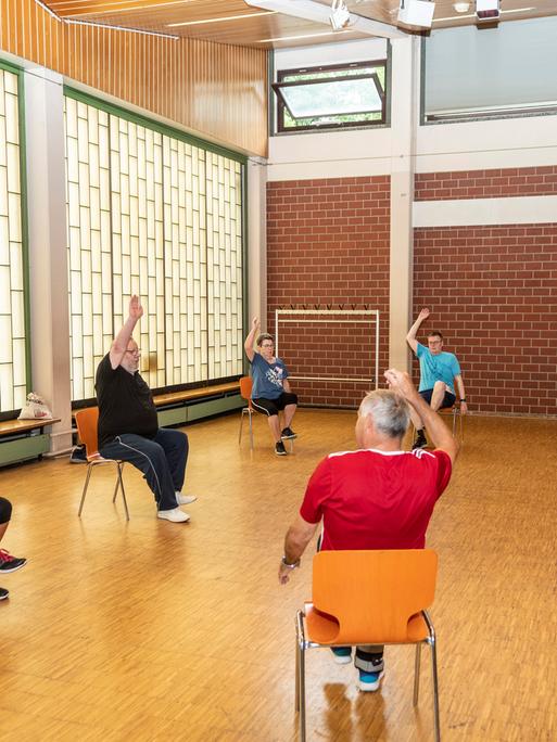 Senioren sitzen  im Rahmen eines Präventionskurses in einer Sporthalle auf Stühlen und strecken einen Arm nach oben. 