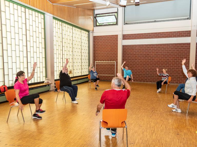 Senioren sitzen  im Rahmen eines Präventionskurses in einer Sporthalle auf Stühlen und strecken einen Arm nach oben. 
