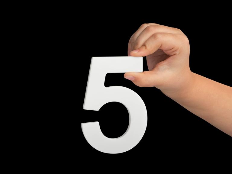 Eine kindliche Hand hält mit spitzen Fingern eine weiße Ziffer 5 vor schwarzem Hintergrund.