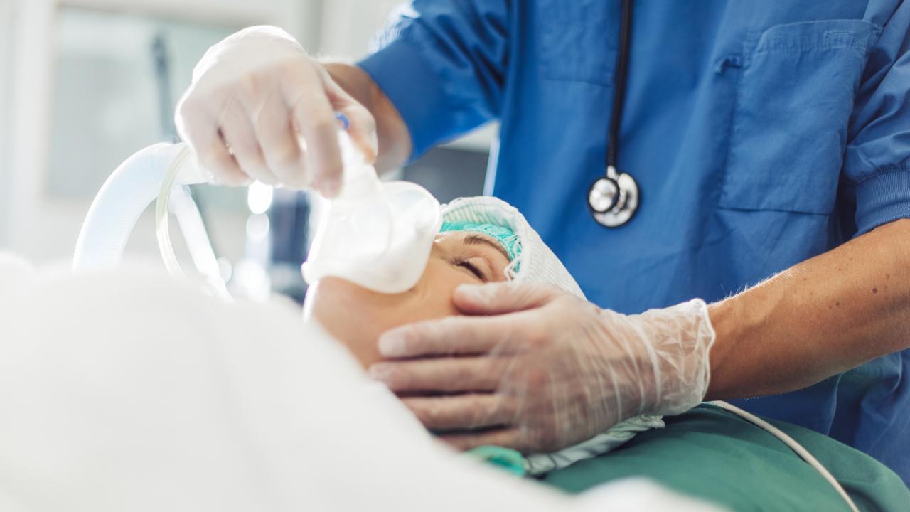 Einer Person mit geschlossenen Augen wird von einer Person aus dem medizinischen Sektor eine Narkosemaske aufgesetzt.