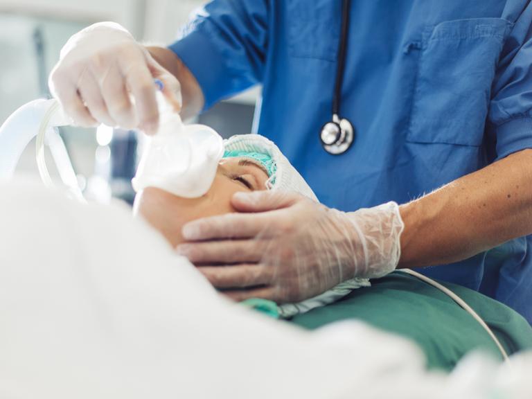 Einer Person mit geschlossenen Augen wird von einer Person aus dem medizinischen Sektor eine Narkosemaske aufgesetzt.