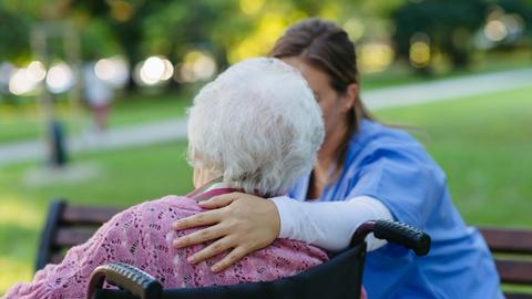 Eine Pflegeperson legt den Arm um eine alte Person. Die alte Person sitzt in einem Rollstuhl und ist lediglich von hinten zu erkennen.