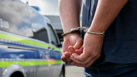 Ein Mann mit Handschellen. Die Hände sind auf dem Rücken gefesselt. Im Hintergrund steht ein Polizeiauto.