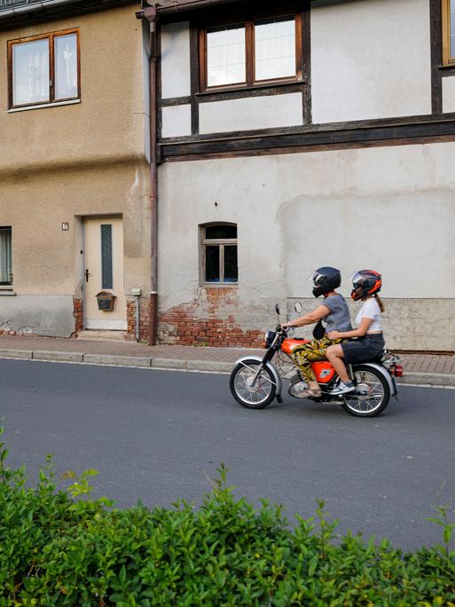 Ein Junge und ein Mädchen fahren auf einem Simson-Moped an alten Häusern in einer Dorfstraße in Thüringen vorbei.