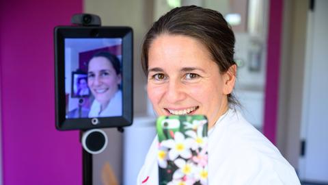 Die Gynäkologin Mandy Mangler hält ein Smartphone in der Hand. Hinter ihr sieht man ihr Gesicht auf einem Monitor.