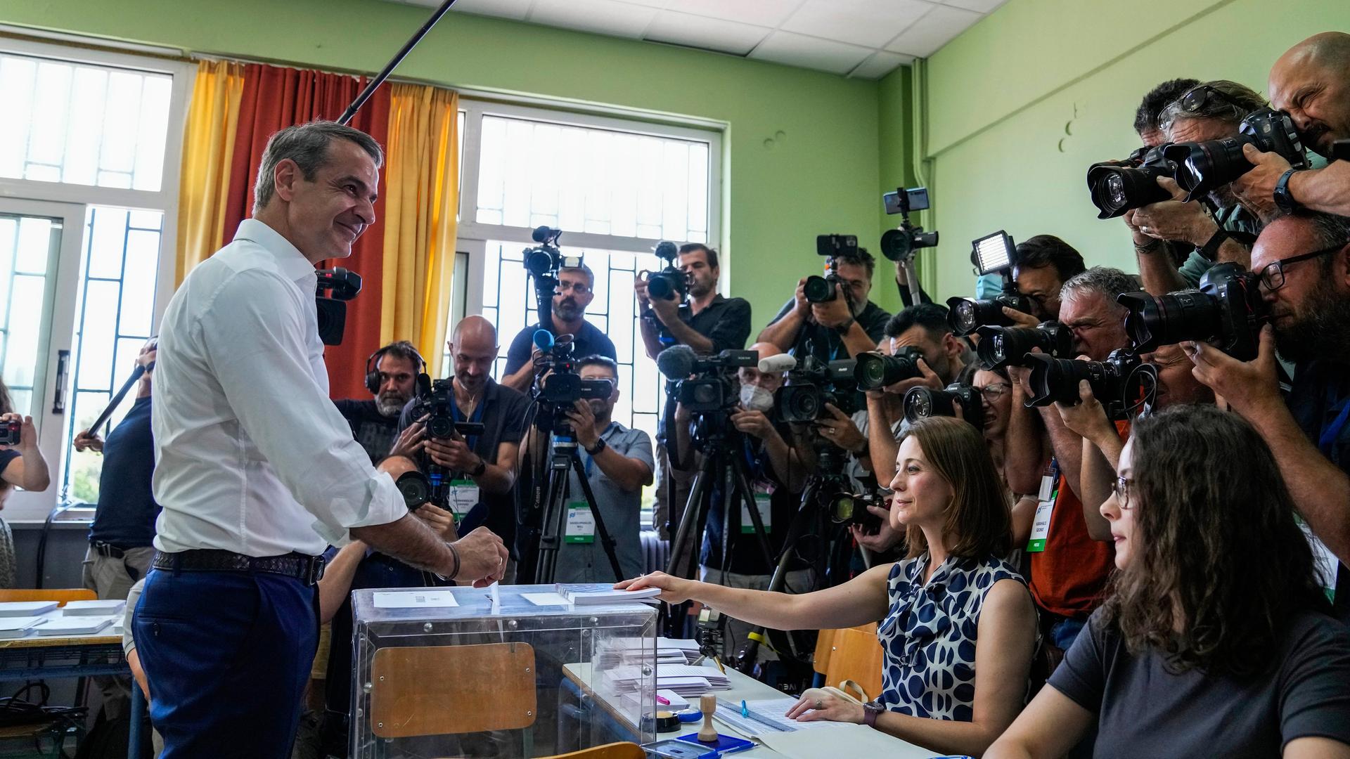 Kyriakos Mitsotakis steckt seinen Stimmzettel in einem Athener Wahllokal in eine Wahlurne. Mit ihm zu sehen sind zwei seitzende Wahlhelferinnen und eine größere Menge an Reportern mit Fotoapparaten, Kameras und Mikrofonen. Kyriakos Mitsotakis steckt seinen Stimmzettel in einem Athener Wahllokal in eine Wahlurne. Mit ihm zu sehen sind zwei seitzende Wahlhelferinnen und eine größere Menge an Reportern mit Fotoapparaten, Kameras und Mikrofonen.