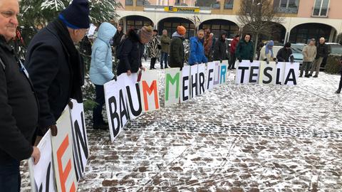 Menschen stehen im Halbkreis auf einem gepflasterten Platz. Sie halten Transparente mit einzelnen Buchstaben vor sich, von links nach rechts kann man lesen: "Kein Baum mehr für Tesla"