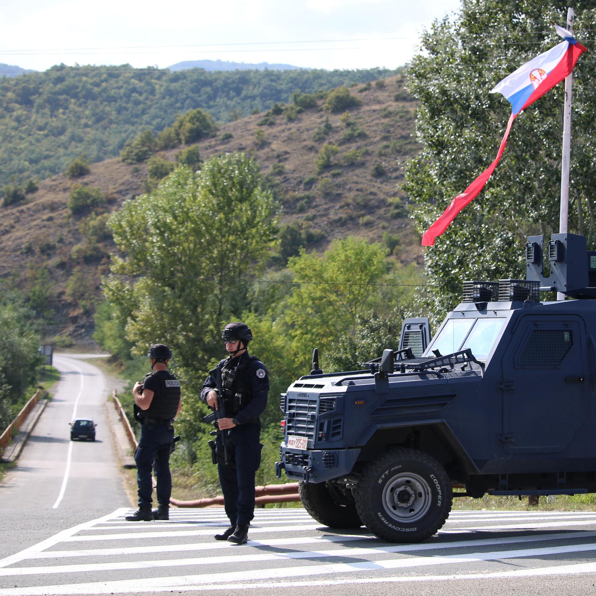 Am Rande einer asphaltierten Strasse in den Bergen steht ein kosovarischer Polizeibeamter und ein Soldat der NATO-Friedensgruppe vor einem Jeep und überwachen die Strasse.
